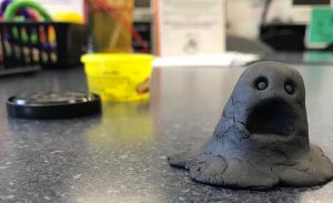 A ghost made out of black Play-Doh in the foreground made to look like it is screaming, with the Play-Doh canister in the background on a writing center table