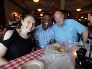 A young man, a young woman and an older man sitting at a restaurant table, smiling into the camera.