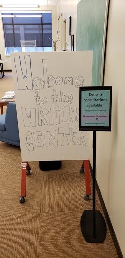 Writing Center entryway. Small sign on the right and in the foreground reads, "Drop in consultations available!" Larger sign to the left and in the background reads, "Welcome to the Writing Center!"