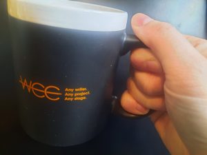 Picture of hand holding a coffee mug, which has the words, "WCC. Any writer. Any Project. Any Stage." written on it.