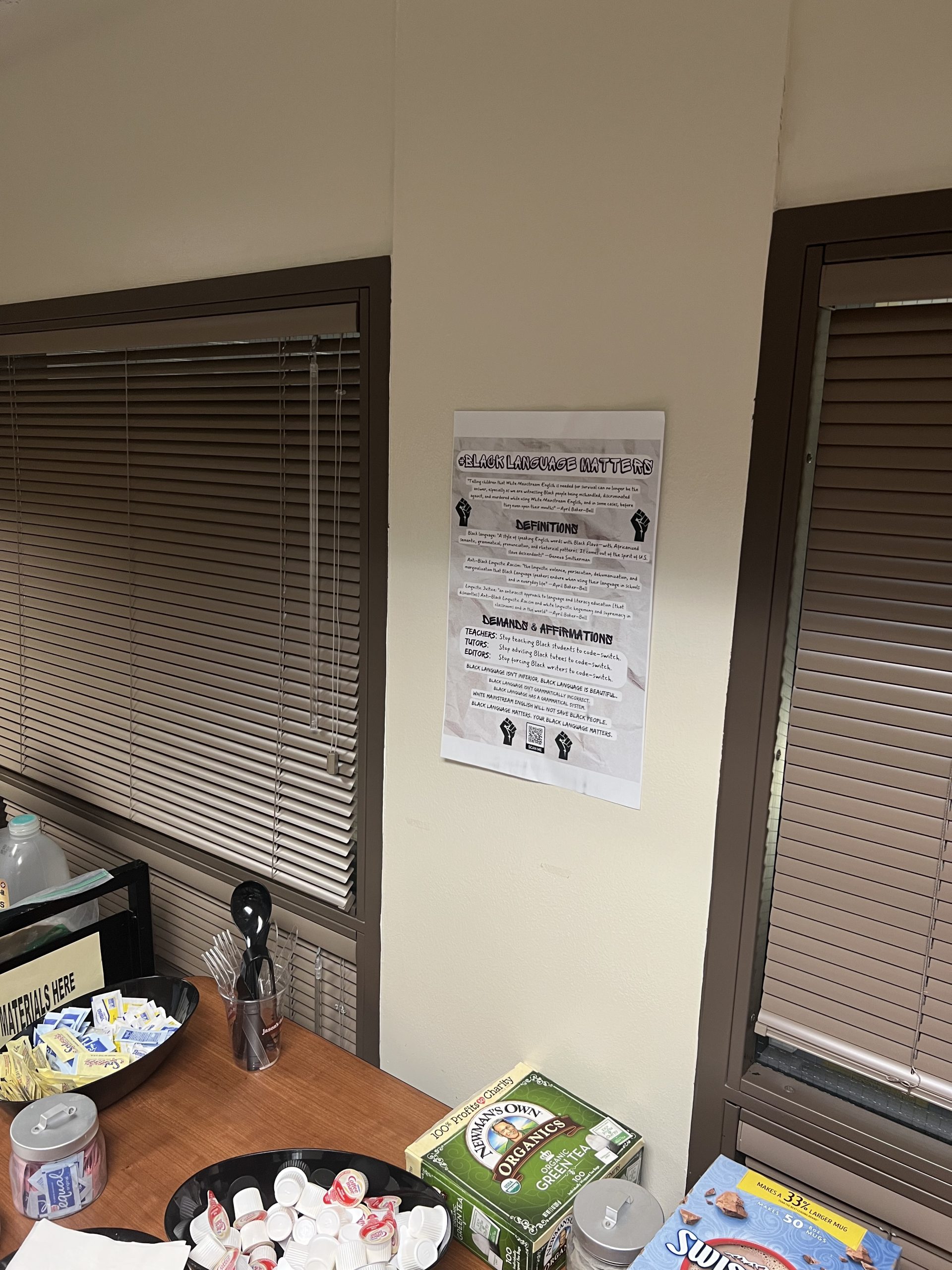 Figure 7: #BlackLanguageMatters poster taped to a wall in between two windows with brown blinds. In front of the poster is a table with various beverages.