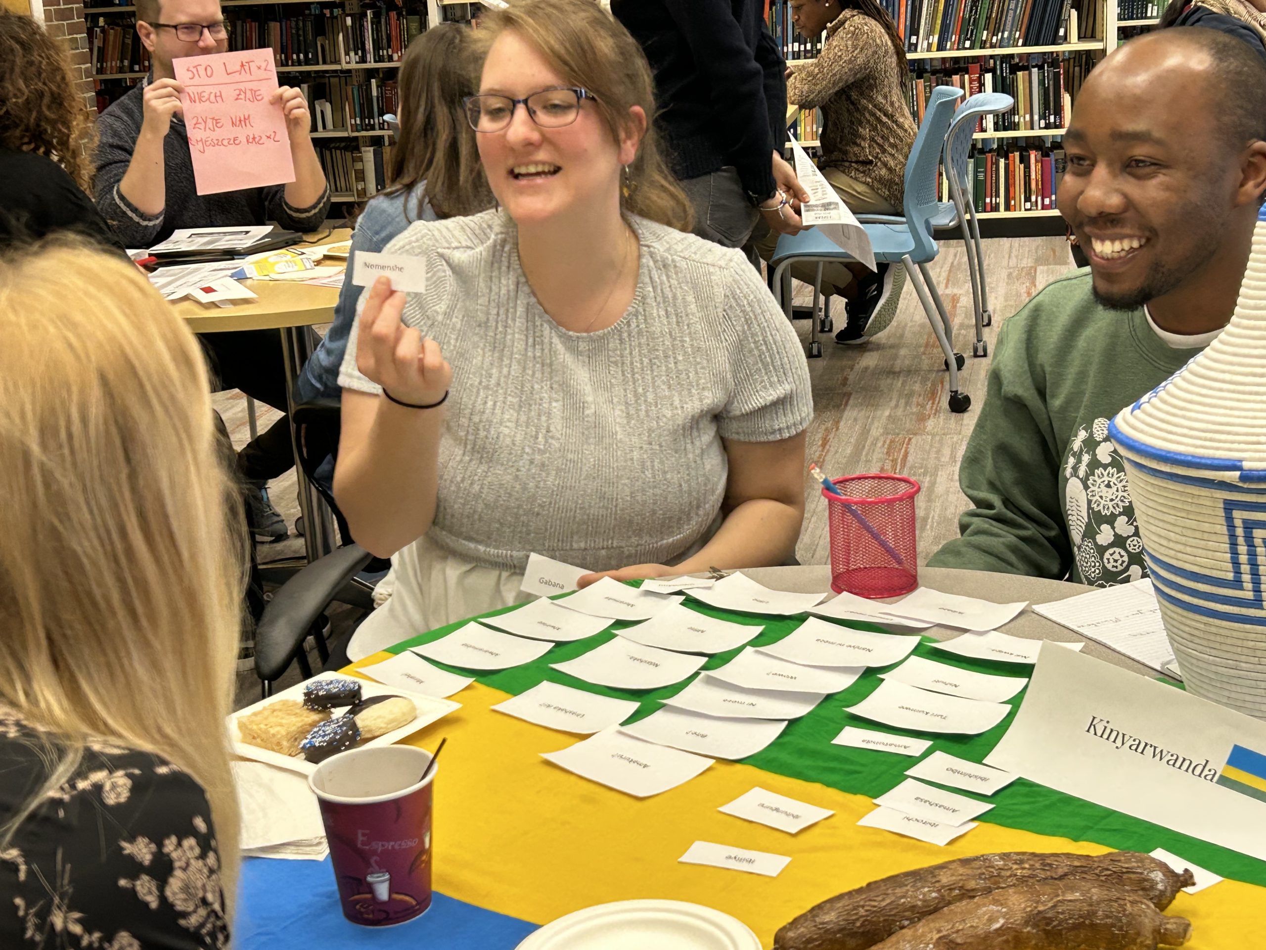 Photo of two individuals, Margaret, co-author of this article and Lucky, writing center visitor, teaching Kinyarwanda. The table where they sit is covered by a Rwandan flag, topped with a sign that says "Kinyarwanda," and flashcards with Kinyarwanda words and phrases.